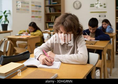 Caucasian boy teenager sitting at desk writing in notebook in classroom with multiethnic classmates studying in background, focused on schoolwork during lesson Stock Photo