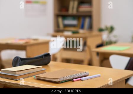 Empty classroom showing wooden desks with notebooks, pencils, and pen case arranged neatly on surface, blurred background with bookshelves and chairs indicating educational environment Stock Photo