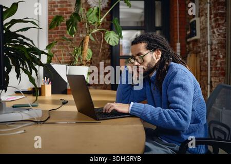 Young Adult Black Man Focusing on Laptop While Sitting at Shared Office Table Stock Photo