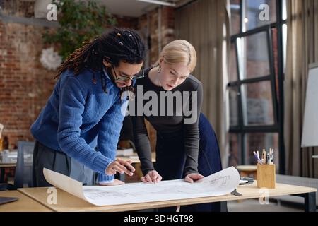 Young Adult Black Man and Caucasian Woman Collaborating on Architectural Plans Stock Photo