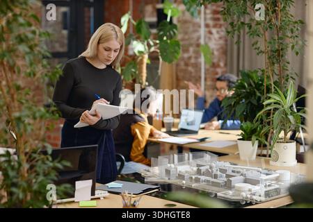 Young Female Architect Writing Notes While Working in Modern Office Workspace Stock Photo