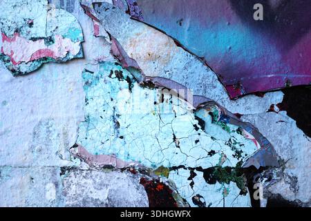 Peeling layers of paint reveal a complex texture on a wall, showcasing vibrant hues of blue, purple, and white. The paint is cracked and flaking, expo Stock Photo