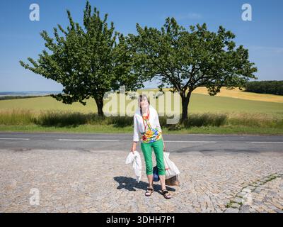 A 50s woman carrying white shopping bags, standing on a cobblestone area by a road with two green trees and a yellow field in the Czech Republic. Stock Photo