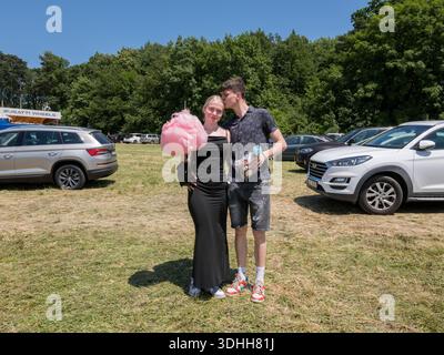 A 20s couple posing at a funfair in the Czech Republic, with the woman holding pink cotton candy while the man kisses her forehead on a grassy lot. Stock Photo