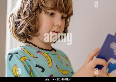 A boy is focused on a book while wearing pajamas. He is in his bedroom and seems engaged in reading, showing interest in the content of the book. Stock Photo