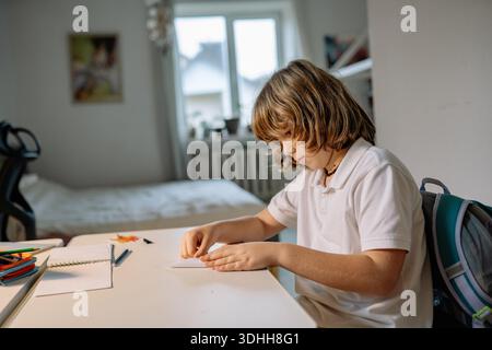 A boy sits at a desk in his room focusing on craft work. He is using paper and tools to create something. Sunlight enters through the window behind hi Stock Photo