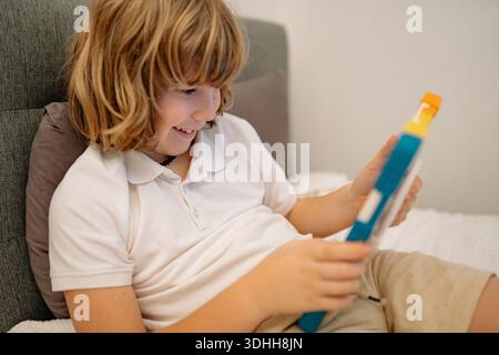 A boy enjoys playing on a tablet while sitting on his bed in his bedroom. He smiles as he interacts with the device, fully engaged in his activity. Stock Photo