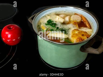 Green pot with cooking water and sliced vegetable, red apple timer, all on black background of ceramic hob. Preparation food. Stock Photo