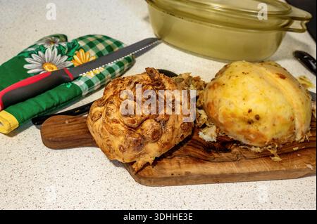 Halved cooked celery root on olive wood cutting board, flower pot holder with knife, glass cooking pan on background, closeup. Preparation of meal. Stock Photo