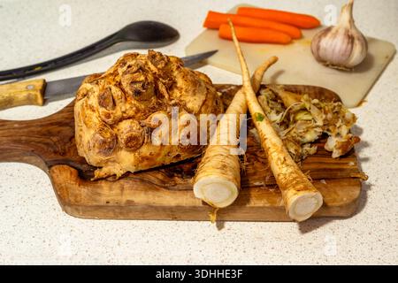 Halved celery root and parsley on olive wood cutting board, carrot and onion, spoon and knife on kitchen table. Preparation of meal. Stock Photo