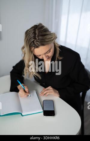 A blonde woman in a black jacket sits at a white table writing in an open notebook. A phone is on the table near the notebook. The activity appears to be taking place in an office setting. Stock Photo
