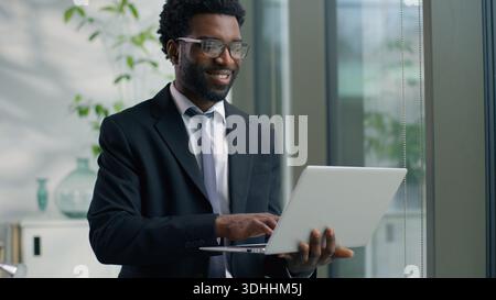 African American businessman male business man in office looking out window typing laptop smiling happy working at workplace using corporate software Stock Photo