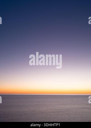 Minimalist seascape shows calm ocean and gradient sky with lavender and warm orange at horizon. Long exposure smooths water. Soft light. Stock Photo