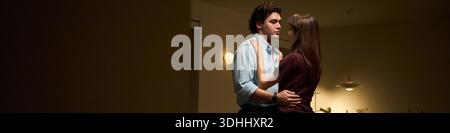 Header of young Caucasian man and young Caucasian woman standing close together embracing in dimly lit room, both looking at each other with serious expressions, intimate moment captured Stock Photo
