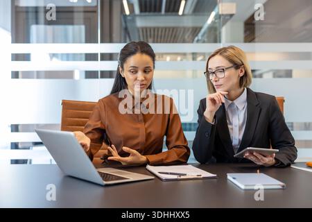 Two smart businesswomen collaborating in a modern office, analyzing data on a laptop and tablet, planning new projects while working together at a conference table Stock Photo