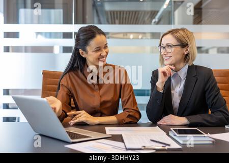 Two smiling multi-ethnic business women discussing a project, collaborating happily in a modern corporate office, using a laptop and documents to analyze data during a professional meeting Stock Photo
