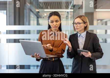 Two professional businesswomen collaborating and discussing a business project in a modern office. One holding a laptop while the other holds a tablet. Symbolizing teamwork and corporate communication Stock Photo