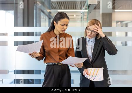 Two diverse businesswomen reviewing documents with concerned and frustrated expressions, indicating financial stress, complex problems, or unexpected challenges in their corporate office environment Stock Photo