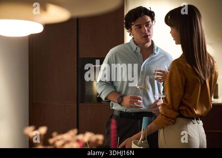 Young Caucasian man talking with young Caucasian woman while holding wine glasses in modern kitchen, both standing close together and engaging in relaxed conversation Stock Photo