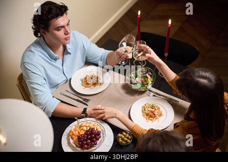 Caucasian young adult man and Caucasian young adult woman sitting at dining table, holding hands and clinking wine glasses, while sharing meal with plates of pasta and salad Stock Photo