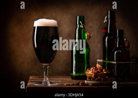 Glass of dark beer with foam head on dark wooden background with empty bottles and beer snacks Stock Photo