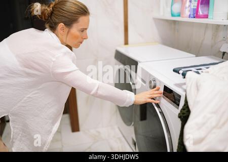 A woman is washing clothes in her laundry room. She is adjusting the settings on the washing machine while standing next to it. Stock Photo