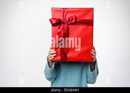 Studio shot of anonymous woman hiding face behind big red gift box with ribbon, isolated on white. Ideal for ecommerce banners, ads, and seasonal prom Stock Photo