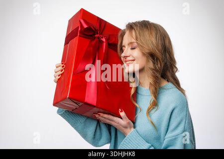 Smiling woman in blue sweater holding big red wrapped gift box on white background, ideal for ecommerce website banners and holiday promotion ads. Stock Photo