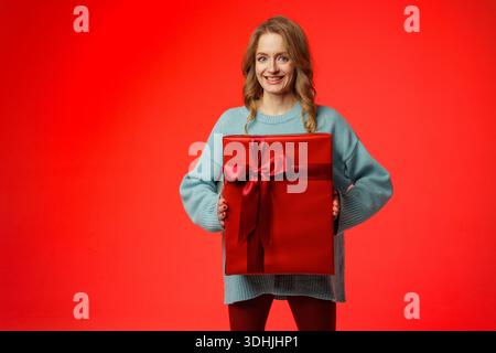 Smiling woman in sweater holding large wrapped gift box on red studio backdrop, ideal for website banner or social ad for holiday promotions. Stock Photo