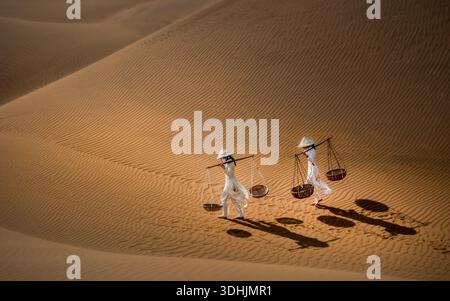 Aerial view of two figures draped in white, carrying baskets, traverse the undulating golden dunes, their shadows a stark contrast on the rippled sand Stock Photo