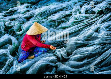 View of a woman in traditional conical hat meticulously mending vast, rippling blue fishing nets under the open sky, Phan Rang, Thap Cham, Vietnam. Stock Photo