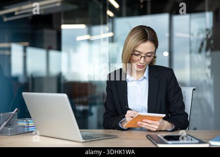 Young businesswoman wearing eyeglasses writing on a notepad while working at a desk with a laptop, showcasing organization, focus, and productivity in a contemporary office environment Stock Photo