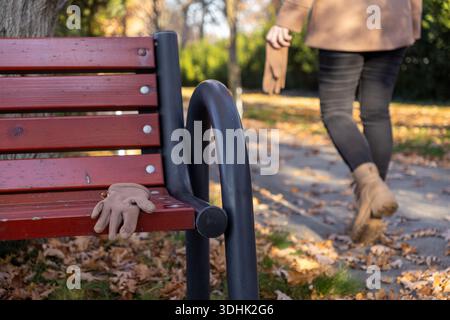Close-up view of a lonely beige glove lying on a reddish-brown slatted wooden bench, with fallen autumn leaves scattered around in a park Stock Photo