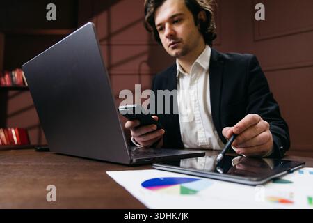 Male professional in a black suit engages with a smartphone and tablet while seated at a wooden desk with business charts and graphs in view Stock Photo