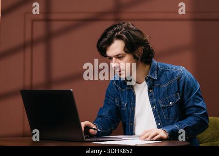 Young man in denim jacket uses laptop on wooden table, displaying a focused expression against a brown wall with soft shadows Stock Photo