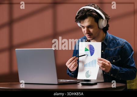 Young man with headphones shows data charts while seated at a table with a laptop, in a contemporary workspace featuring a brown wall Stock Photo