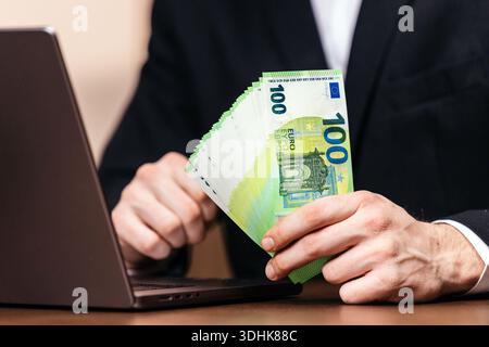 Individual in formal attire displays a bundle of 100 euro notes while working on a laptop at a wooden desk in an office environment Stock Photo