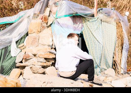 Person in white hoodie rests on rocks beside a rudimentary shelter made of stones and patterned materials outdoors. Stock Photo
