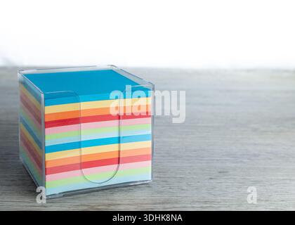 A clear acrylic cube filled with a stack of vibrant, multi-colored square memo papers on a wood desk. Stock Photo