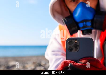Individual wearing blue gas mask and red gloves holding smartphone on pebble beach by the sea. Stock Photo