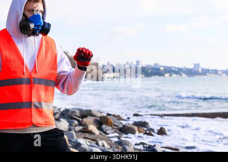 Individual in safety attire uses mobile on rocky beach with sea and urban horizon in view Stock Photo