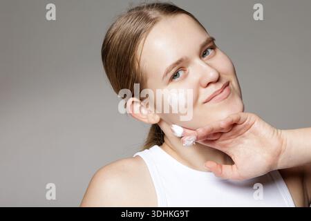 Studio portrait of young woman applying moisturizer with heart cream on cheek. Suitable for skincare website banner or social ads about daily routine Stock Photo