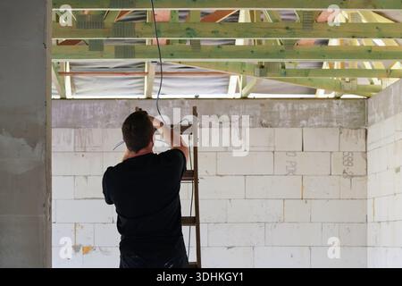 Unrecognizable man standing on a ladder, view from behind - electrician - installing cables at newly built house wall near roof Stock Photo