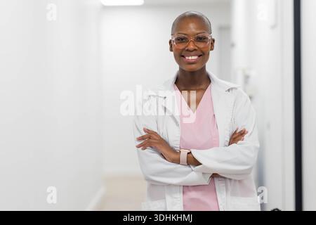 African woman standing in clinic wearing white lab coat, pink scrubs, clear glasses and smartwatch Stock Photo