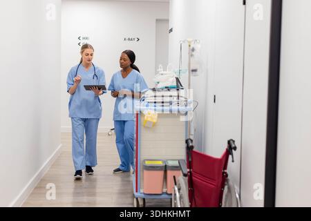 Diverse female nurses walking down hospital corridor in light blue scrubs, one pointing at tablet Stock Photo