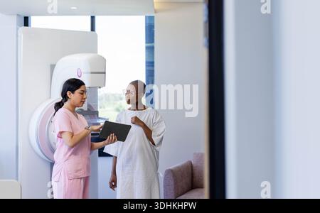 Diverse female tech in pink scrubs consulting patient in gown at mammography machine, using tablet Stock Photo