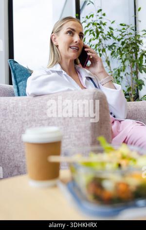 Female clinician sitting on sofa in clinic speaking on phone, wearing lab coat and stethoscope Stock Photo