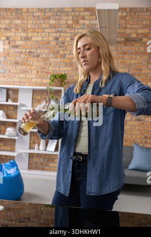 Woman wearing denim shirt pouring beer from green bottle into pilsner glass in loft living area Stock Photo