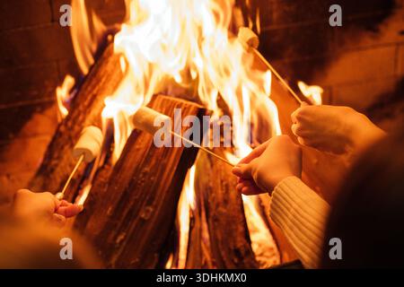 Hands are holding skewers with marshmallows roasting over a warm fire, glowing flames and logs create a cozy atmosphere for gatherings Stock Photo