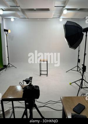 Empty photography studio setup with lights, softbox, camera on tripod and stool on white backdrop. Stock Photo